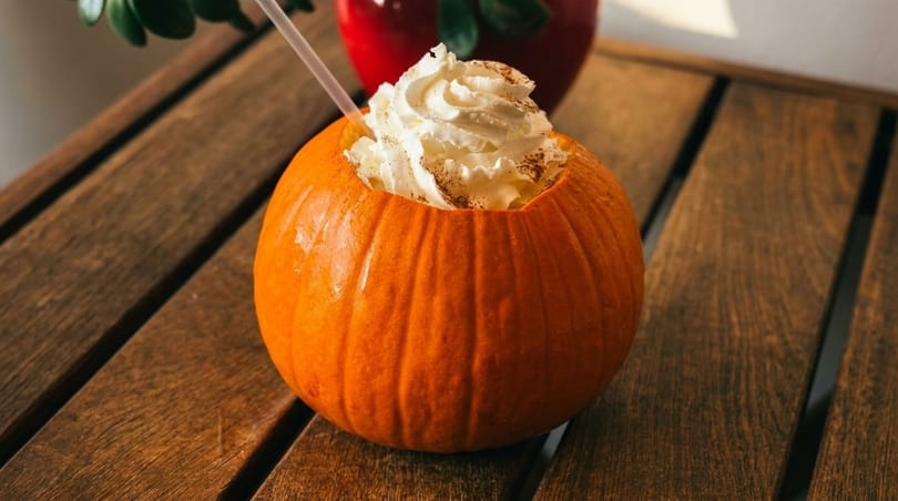 Pumpkin drink with whipped cream and cinnamon on a wooden table, with a red pot in the background.